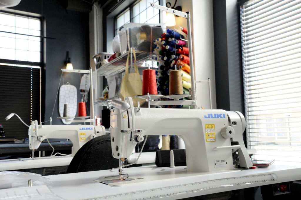 Photo of a Juki industrial sewing machine in a sunlit sewing studio. A vertical rack of colorful thread spools hangs in the background.